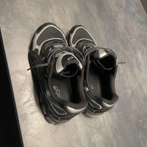 Svarta och grå sneakers från Asics - Säljer ett par snygga och bekväma svarta och grå sneakers från Asics. Skorna har en sportig design med snörning och är perfekta för träning eller vardagsbruk. De har en rund tå och är tillverkade i syntetmaterial för extra hållbarhet. Dem är bara andvända 2 gånger så dem är i princip ny skick, priset kan sänkas vid snabb äffar. Ingen box kommer. 
