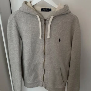 Polo Ralph Lauren grå hoodie storlek M - Grå zip-up hoodie från Polo Ralph Lauren. Klassisk modell med dragkedja fram och luva. Perfekt för en avslappnad men stilfull look.