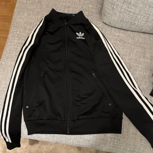 Svart träningsjacka från Adidas - Snygg svart träningsjacka från Adidas med klassiska vita ränder längs ärmarna och logga på bröstet och ryggen. Jackan har dragkedja framtill och två fickor med dragkedjor. Perfekt för en sportig look! Storlek: 34 