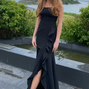 Svart långklänning med volangdetaljer - Elegant svart långklänning med smala axelband och volangdetaljer framtill. Klänningen har en öppen rygg med snörning som ger en stilren look. Perfekt till bal. Endast använd 1 gång💕