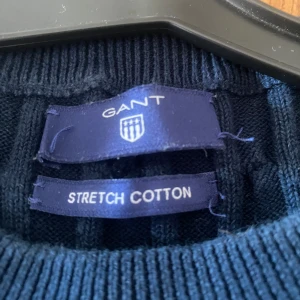 Mörkblå kabelstickad tröja från GANT - Säljer en stilren mörkblå kabelstickad tröja från GANT i stretchig bomull. Tröjan har en klassisk rund halsringning och GANT-loggan broderad på bröstet. 