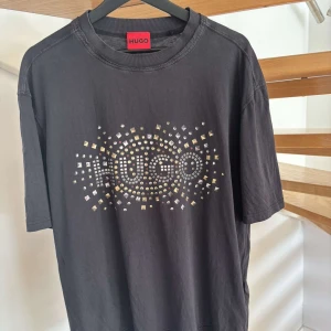Svart t-shirt från Hugo - Snygg svart t-shirt från Hugo med ett coolt mönster av små fyrkanter och texten 'Hugo' i mitten. Perfekt för en stilren look med en touch av bling. Kortärmad och i mjukt material.