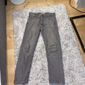Grå jeans från Jack & Jones - Säljer ett par grå jeans från Jack & Jones, modell Relaxed/Chris. De har en avslappnad passform och är perfekta för en casual look. Jeansen har en klassisk femficksdesign och stängs med knapp och dragkedja.