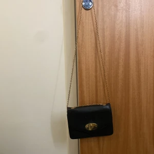 Svart axelväska med kedja - Snygg svart axelväska med guldfärgad kedja och ett elegant spänne framtill. Perfekt för att bära det nödvändigaste med stil. Väskan har en praktisk dragkedja inuti för extra säkerhet.