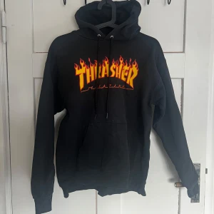 Svart hoodie från Thrasher - Säljer en svart hoodie från Thrasher med det ikoniska flamtrycket i gult och rött på bröstet. Tröjan har en stor ficka framtill och justerbar huva med snören. Perfekt för en avslappnad stil.