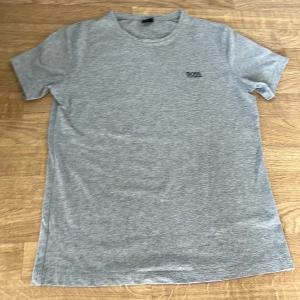 Grå t-shirt från Hugo Boss - Säljer en stilren grå t-shirt från Hugo Boss med broderad logga på bröstet. Perfekt för en avslappnad look. T-shirten är kortärmad och gjord i mjukt material.