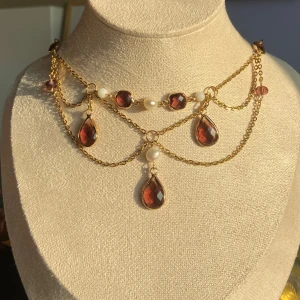 Guldhalsband med pärlor och röda/lila stenar - Elegant guldhalsband med hängande röda droppformade stenar och vita pärlor. Kedjan har flera lager och små röda stenar som ger en lyxig känsla. Perfekt för att lyfta vilken outfit som helst.
