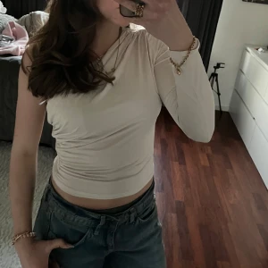Beige oneshoulder topp - Säljer en stilren beige oneshoulder topp med lång ärm. Toppen har en snygg rynkad detalj på axeln och är perfekt för en trendig look. Passar bra till både jeans och kjol. Perfekt för att uppgradera garderoben!