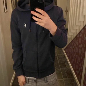 Mörkblå hoodie från Ralph Lauren - Säljer en snygg mörkblå hoodie från Ralph Lauren med dragkedja och klassisk logga på bröstet. Perfekt för en avslappnad stil. Den har långa ärmar och en praktisk huva med snören den är dock hyfsat solblekt. 