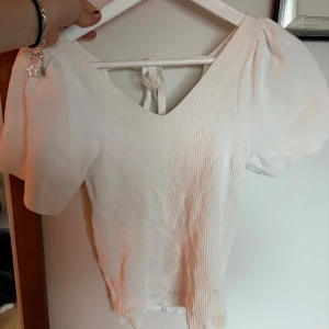 Vit ribbad topp från ONLY - Säljer en stilren vit ribbad topp från ONLY med v-ringning och korta puffärmar. Toppen har en knytning i ryggen som ger en fin detalj. Perfekt för en somrig look! 🌼