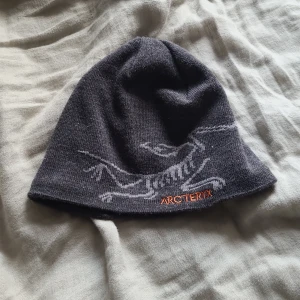 Grå mössa från Arc'teryx - Snygg grå mössa från Arc'teryx med ett coolt mönster och orange broderad logga. Perfekt för kyliga dagar.