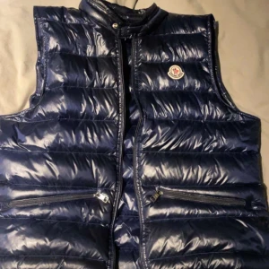 Blå dunväst från Moncler - Snygg blå dunväst från Moncler med glansig finish. Västen har dragkedja och två praktiska fickor med dragkedjor. Perfekt nu till våren!