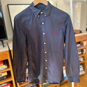 Mörkblå skjorta från Ralph Lauren - Säljer en stilren mörkblå skjorta från Ralph Lauren i slim fit. Skjortan har långa ärmar och knappar framtill. Den är tillverkad i bomull och har en broderad logotyp på bröstet. Perfekt för en klassisk look! Storlek 170 och motsvarar typ XS herr eller S dam.