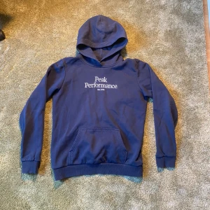 Peak Performance Hoodie - Nypris: 600 kronor Säljer en snygg mörkblå hoodie från Peak Performance med vit logga på bröstet. Tröjan har en klassisk känguruficka och en bekväm huva. Perfekt för en avslappnad stil.