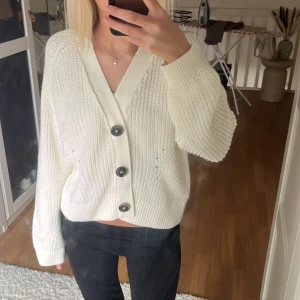 Vit stickad kofta med knappar - Säljer en mysig vit stickad kofta med stora knappar framtill. Koftan har en v-ringad design och långa ärmar, perfekt för kyligare dagar. Den är både stilren och bekväm.