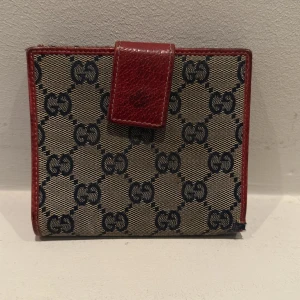 Mönstrad plånbok från Gucci - Säljer en elegant plånbok från Gucci med klassiskt mönster i beige och rött . Plånboken har röda skinnkanter och en praktisk knappstängning. Insidan är röd med flera kortfack för smidig organisering. Finns även fack för mynt år rfekt för den stilmedvetna!