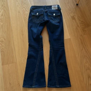 Mörkblå bootcut jeans från True Religion strl 26 - Snygga mörkblå jeans från True Religion med bootcut-stil. De har dekorativa sömmar och knappar på bakfickorna. Perfekta för en avslappnad look.