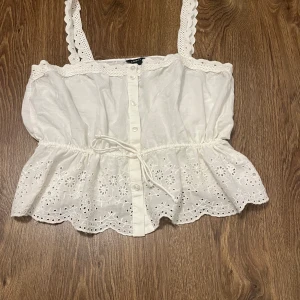 Vit blus från Lindex - Säljer en söt vit blus från Lindex med spetsdetaljer och knappar framtill. Blusen har broderade blommor och en knytning i midjan för en feminin look. Perfekt för varma dagar!