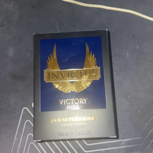 Invictus Victory Elixir Parfum Intense - Säljer en exklusiv Invictus Victory Elixir Parfum Intense från Paco Rabanne. Flaskan är formad som en trofé i en elegant mörkblå färg med en silverfärgad 'V' på framsidan. Parfymen är vegansk och tillverkad i Frankrike, känd för sin långvariga doft. Ungefär 65% kvar av parfymen.