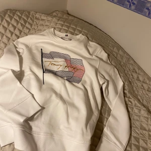 Vit sweatshirt från Tommy Hilfiger - Säljer en stilren vit sweatshirt från Tommy Hilfiger med ett stort flaggmotiv och broderad logga i guld på framsidan. Tröjan har långa ärmar och en klassisk rund halsringning. Perfekt för en avslappnad look. Helt ny och oanvänd med tags kvar!
