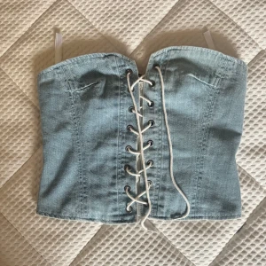 Ljusblå korsettopp med snörning - Trendig ljusblå korsettopp i jeansmaterial. Toppen har en snygg snörning framtill och dragkedja bak för enkel på- och avtagning. Perfekt för att ge en edgy touch till din outfit!