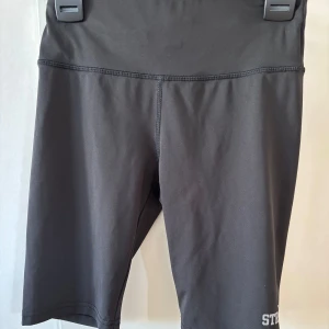 Svarta cykelshorts från OTP Young  - Snygga svarta cykelshorts i syntetmaterial. Men text ”STRONG” på sidan. Perfekta för träning med en tight passform och hög midja. Stilrena och enkla att matcha med olika toppar.