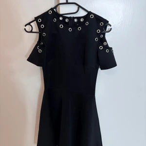 Svart klänning med nitar - Snygg svart klänning med coola nitar runt halsen och ärmarna. Klänningen har en modern offshoulder-design och korta ärmar. Perfekt för en kväll ute eller en speciell tillställning.
