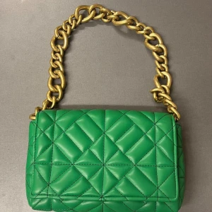 Grön quiltad handväska med kedja - Snygg grön handväska med quiltat mönster och en rejäl guldfärgad kedja som axelrem. Väskan har en stilren design med ett praktiskt innerfack. Perfekt för att ge din outfit en färgklick! Nyskick, aldrig använd!