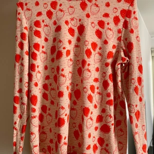 Beige och röd långärmad tröja med jordgubbsmönster - Säljer en beige långärmad tröja från Carin Wester med ett lekfullt rött jordgubbsmönster. Perfekt för att liva upp garderoben med något unikt och färgglatt. Tröjan har en rund halsringning och är tillverkad i ett mjukt material.