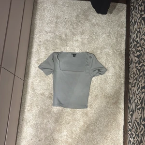 Grå kortärmad topp - Säljer en stilren grå topp med korta ärmar och en fyrkantig halsringning. Perfekt för en minimalistisk look. Passar bra till både jeans och kjol.