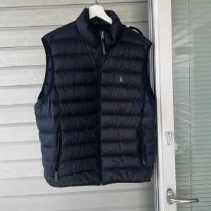 Svart dunväst från Polo Ralph Lauren - Snygg svart dunväst från Polo Ralph Lauren med dragkedja och diskret logga på bröstet. Perfekt för lager-på-lager under kyliga dagar. Västen har en quiltad design och praktiska fickor med dragkedja.
