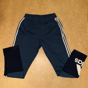 Blå mjukisbyxor från Adidas - Säljer ett par blå mjukisbyxor från Adidas med vita ränder längs sidorna. Byxorna har en elastisk midja med snörning och en logga på benet. Perfekta för en avslappnad stil.