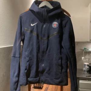 Mörkblå Nike Tech med PSG-logga - Säljer en mörkblå Nike Tech  med PSG-logga på bröstet. Den är köpt 2021 i Paris Saint Germains officiella affär. Den är bra skick men lite slitningar som ni ser på sista bilden! Hör av er om ni har någon fråga angående tröjan, priset kan diskuteras😄