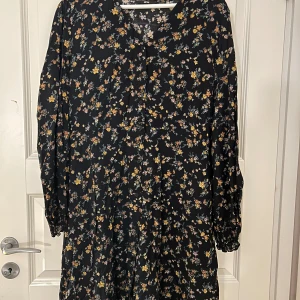 Blommig klänning med knappar - Säljer en svart klänning med ett fint blommigt mönster i gult och blått. Klänningen har långa ärmar och knappar framtill. Svart underklänning hör till. Aldrig använd.