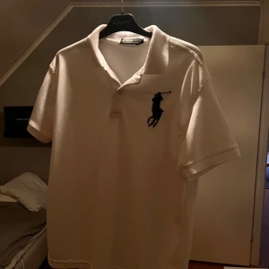 Polo Ralph lauren pike - Säljer en klassisk vit pikétröja från Ralph Lauren med en broderad logga i svart på bröstet. Tröjan har korta ärmar och en traditionell krage med knappar. Perfekt för en stilren och avslappnad look. 