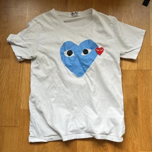 Vit t-shirt från Comme des Garçons - Säljer en vit t-shirt från Comme des Garçons med ett stort blått hjärta och ett litet rött hjärta på framsidan. Perfekt för en stilren och lekfull look. 💙❤️