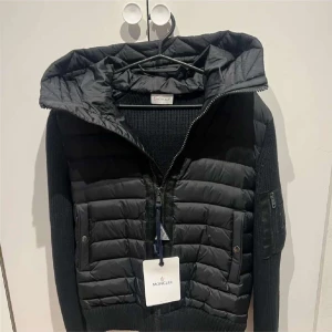 Svart jacka från Moncler - Snygg svart jacka från Moncler med quiltad design och dragkedja. Jackan har en huva och långa ärmar med en praktisk ficka på ärmen. Perfekt för kyligare dagar.