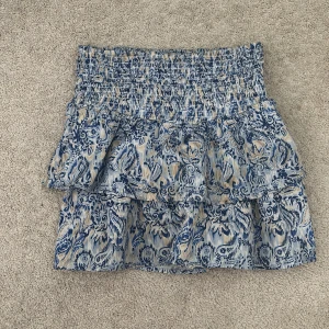 Mönstrad kjol från Gina Tricot - Säljer en snygg kortkjol från Gina Tricot med blått och beige mönster. Kjolen har en volangdetalj och en elastisk midja för en bekväm passform. Perfekt för sommarens alla tillfällen!