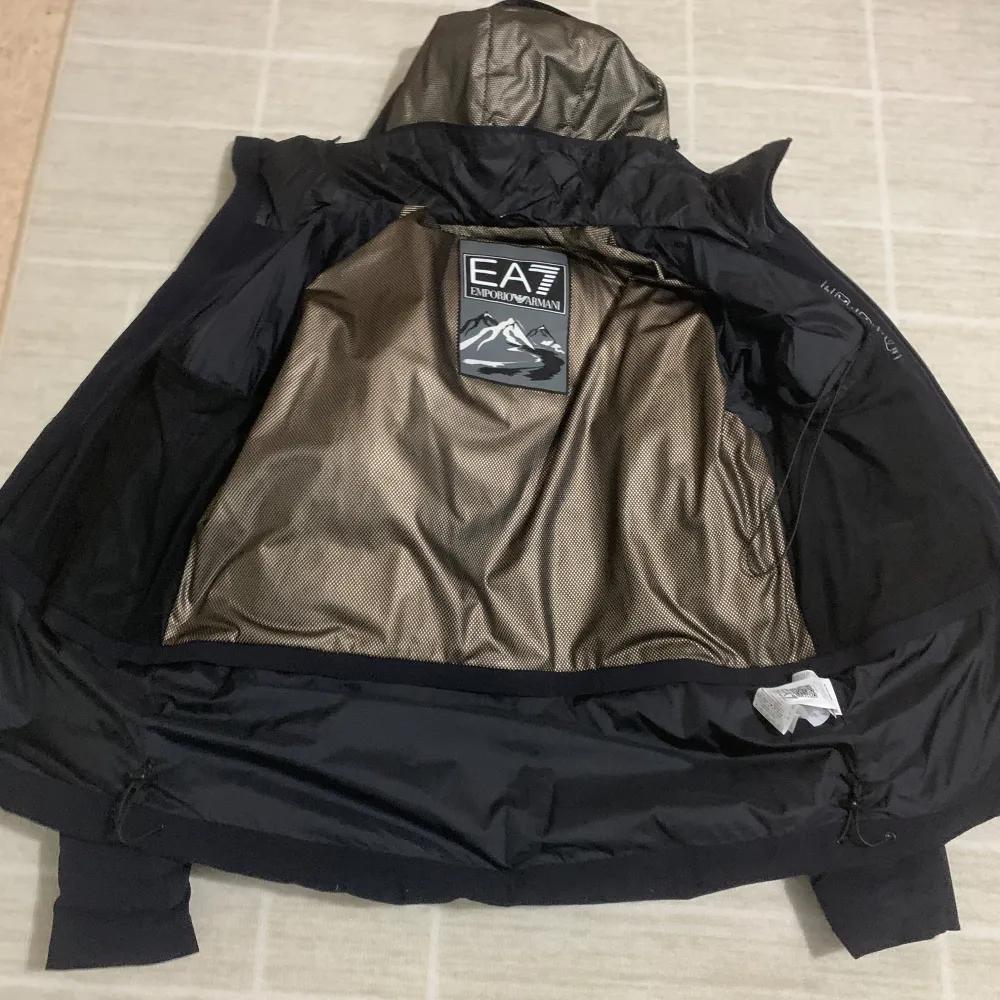 Snygg svart dunjacka från Emporio Armani med huva och dragkedja. Jackan har en stilren design med EA7-logga på ärmen och praktiska fickor med dragkedjor. Perfekt för kyliga dagar. Den har en justerbar luva.. Takit.