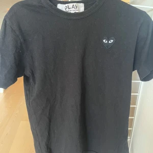 Svart t-shirt från Comme des Garçons - Säljer en svart t-shirt från Comme des Garçons med det ikoniska hjärta med ögon broderat på bröstet. Perfekt för en stilren look. Kortärmad och tillverkad i mjukt material.
