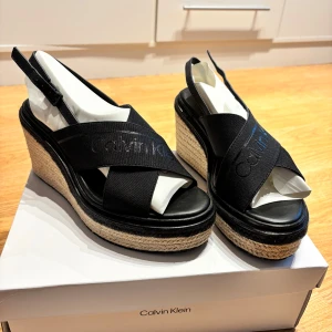 Svarta kilklackar från Calvin Klein - Snygga svarta kilklackar från Calvin Klein med öppen tå och elastiska band. Bara använda en gång. Nypris 1799 kr.