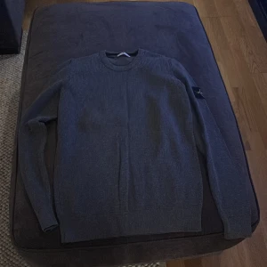 Grå stickad tröja från Stone Island - Säljer en stilren grå stickad tröja från Stone Island. Tröjan har långa ärmar och en rund halsringning. Den ikoniska Stone Island-loggan sitter på ärmen. Perfekt för en avslappnad och trendig look.