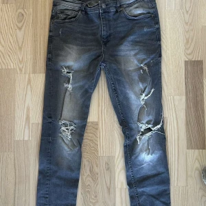 Grå jeans med slitningar från FSBN - Säljer ett par grå jeans från FSBN med coola slitningar på benen. De har en klassisk femficksdesign och knappgylf. Perfekta för en avslappnad stil.