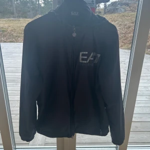 Svart jacka från Emporio Armani EA7 - Snygg svart jacka från Emporio Armani EA7 med dragkedja och huva. Perfekt för en stilren look. Jackan har en diskret logga på bröstet och är tillverkad i ett syntetmaterial. Super fint skick. 