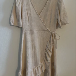 Beige omlottklänning med volang - Säljer en beige omlottklänning med v-ringning och knytband i midjan. Klänningen har trekvartsärmar och en fin volangdetalj längs kanten. Perfekt till sommaren!☀️🩷(aldrig använd)