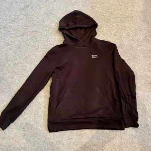 Svart hoodie med tryck Från Kappahl  - Säljer en stilren svart hoodie med en liten vit text på bröstet. Den har en klassisk känguruficka och en bekväm huva. Perfekt för en avslappnad look.