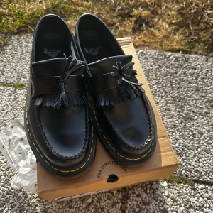 Svarta loafers från Dr. Martens - Snygga svarta loafers från Dr. Martens med klassisk tofsdetalj och fransar. Skorna har en robust sula med den ikoniska gula sömmen. Perfekta för en stilren look.