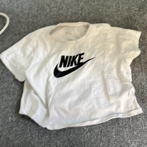 Vit croppad tröja från Nike 146-156 cm - Säljer en vit croppad tröja från Nike med deras ikoniska logga i svart på framsidan. Perfekt för en sportig look eller avslappnad stil. T-shirten är kortärmad och gjord i bomull för en skön känsla.