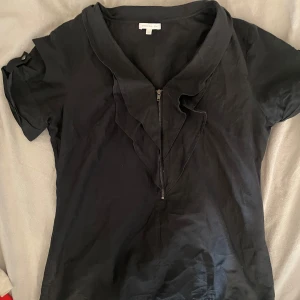 Svart blus med dragkedja  - Snygg svart blus  med korta ärmar och en dragkedja framtill. Blusen har en elegant volangdetalj vid halsen som ger en stilren look. Perfekt för att klä upp en vardagsoutfit eller för en kväll ute.