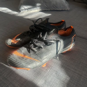 Svarta och orange fotbollsskor från Nike - Säljer ett sällsynt par svarta och orange fotbollsskor från Nike Mercurial. Svåra att hitta. Skorna har en snygg design med snörning och är perfekta för fotbollsplanen. De har en slitstark sula med dobbar för optimalt grepp.